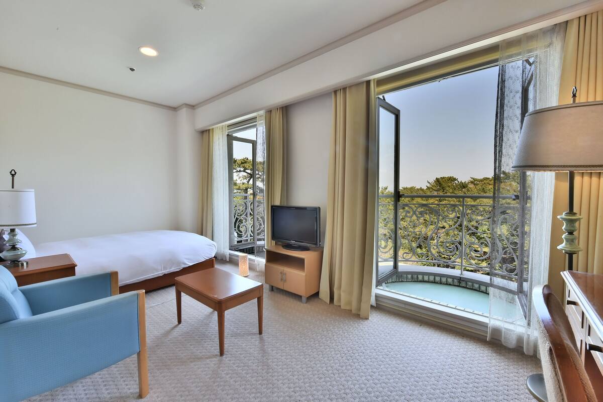 Habitación de hotel con cama, televisión, escritorio, silla y un amplio ventanal con vistas al balcón.
