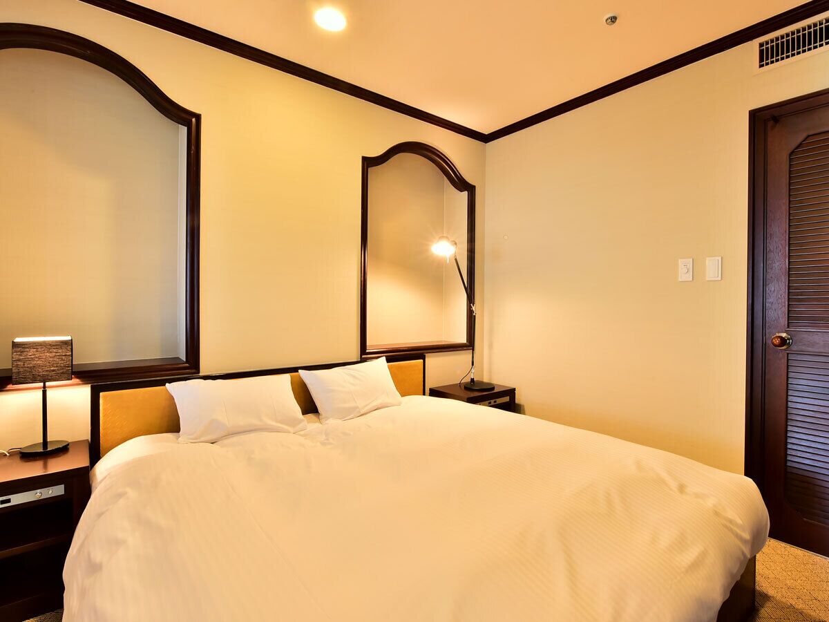 A hotel room with a large bed, a nightstand with a lamp, a mirror, and a door.