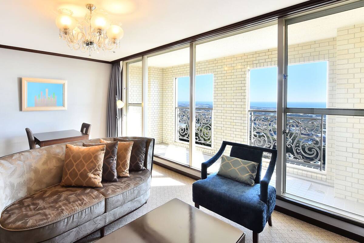 Un salón con sofá, sillón y una mesa. Hay un ventanal con vistas a un paisaje urbano y un balcón con barandilla.