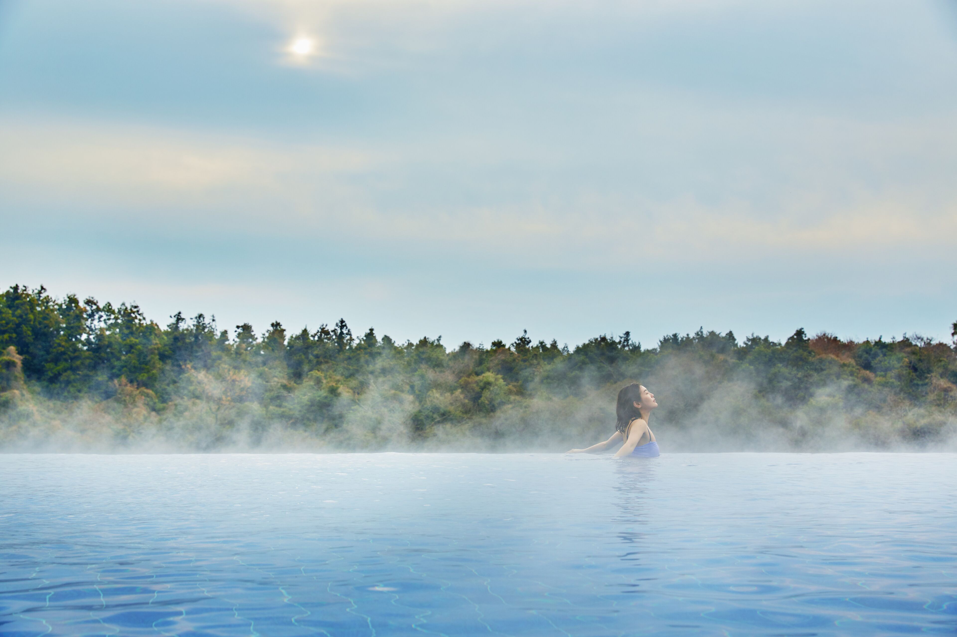 A person in a large, clear pool with steam rising from the water, surrounded by a forested area.