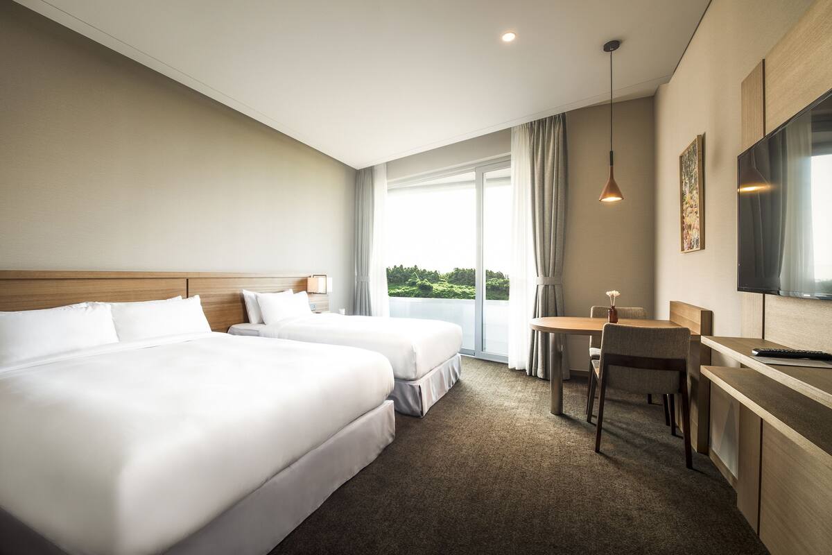 A hotel room with two beds, a desk, a chair, a TV, and a large window with a view of greenery.