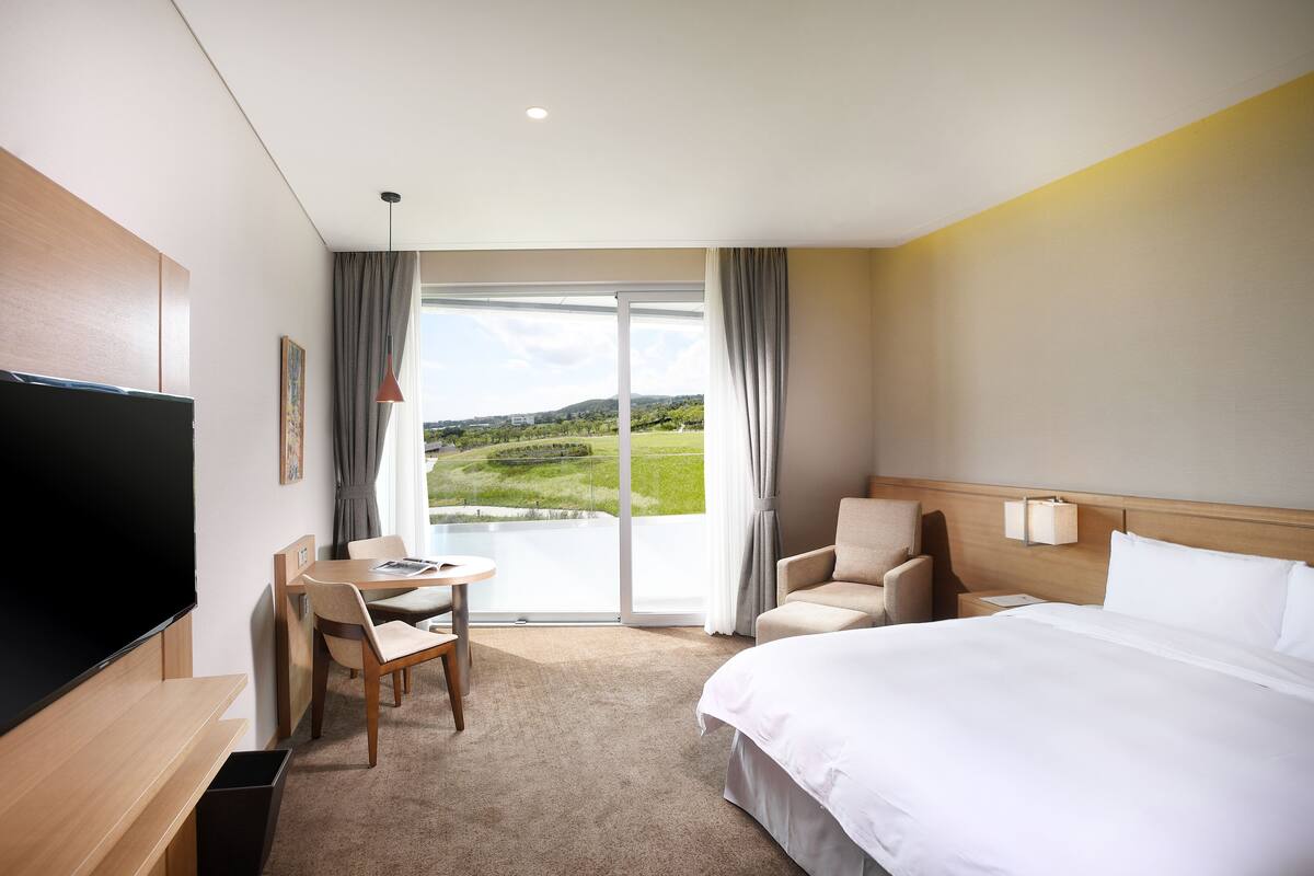 A hotel room with a bed, a desk with a chair, a TV, and a view of a green landscape through a large window.