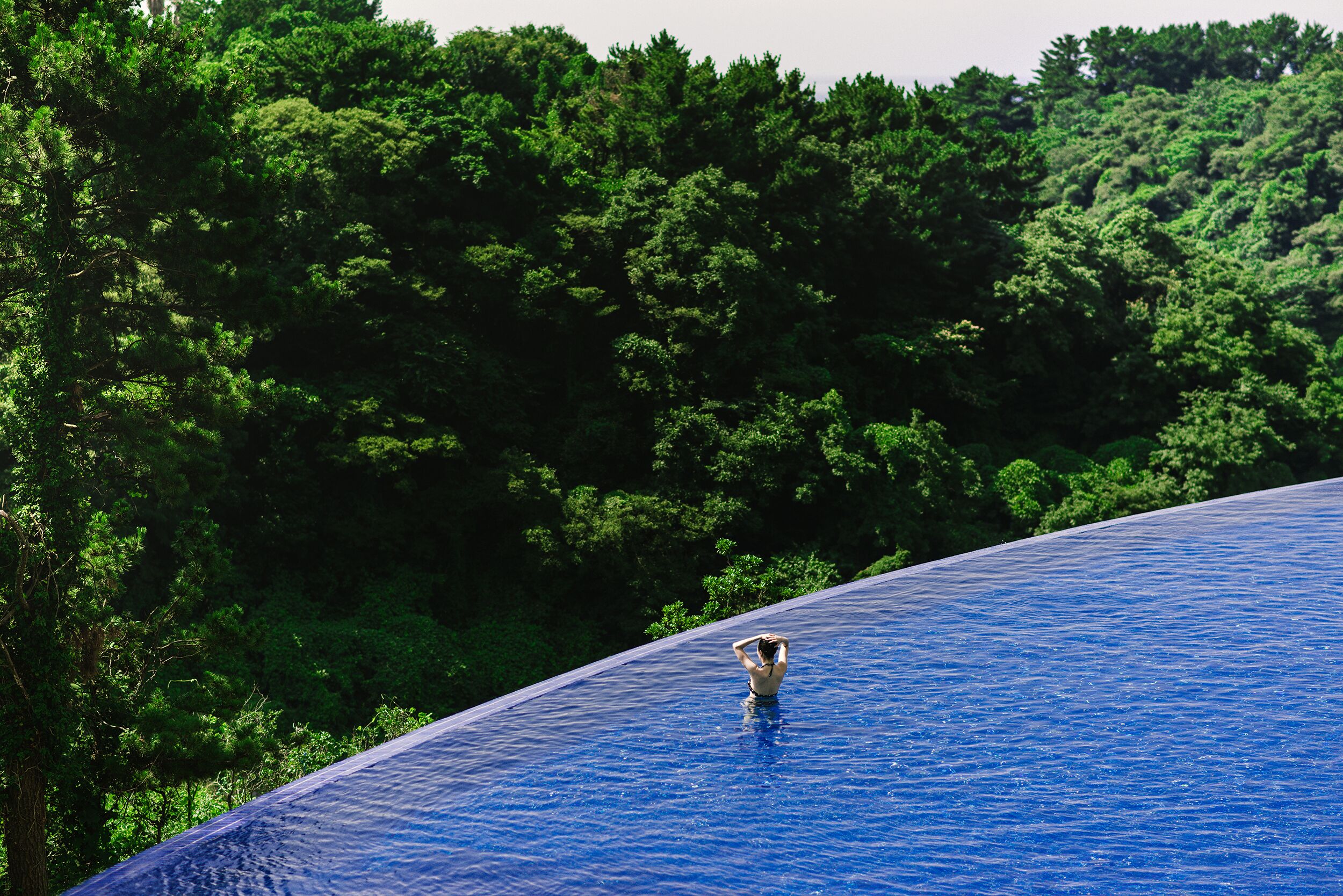 A person standing in a large, clear blue pool with a forested hillside in the background.