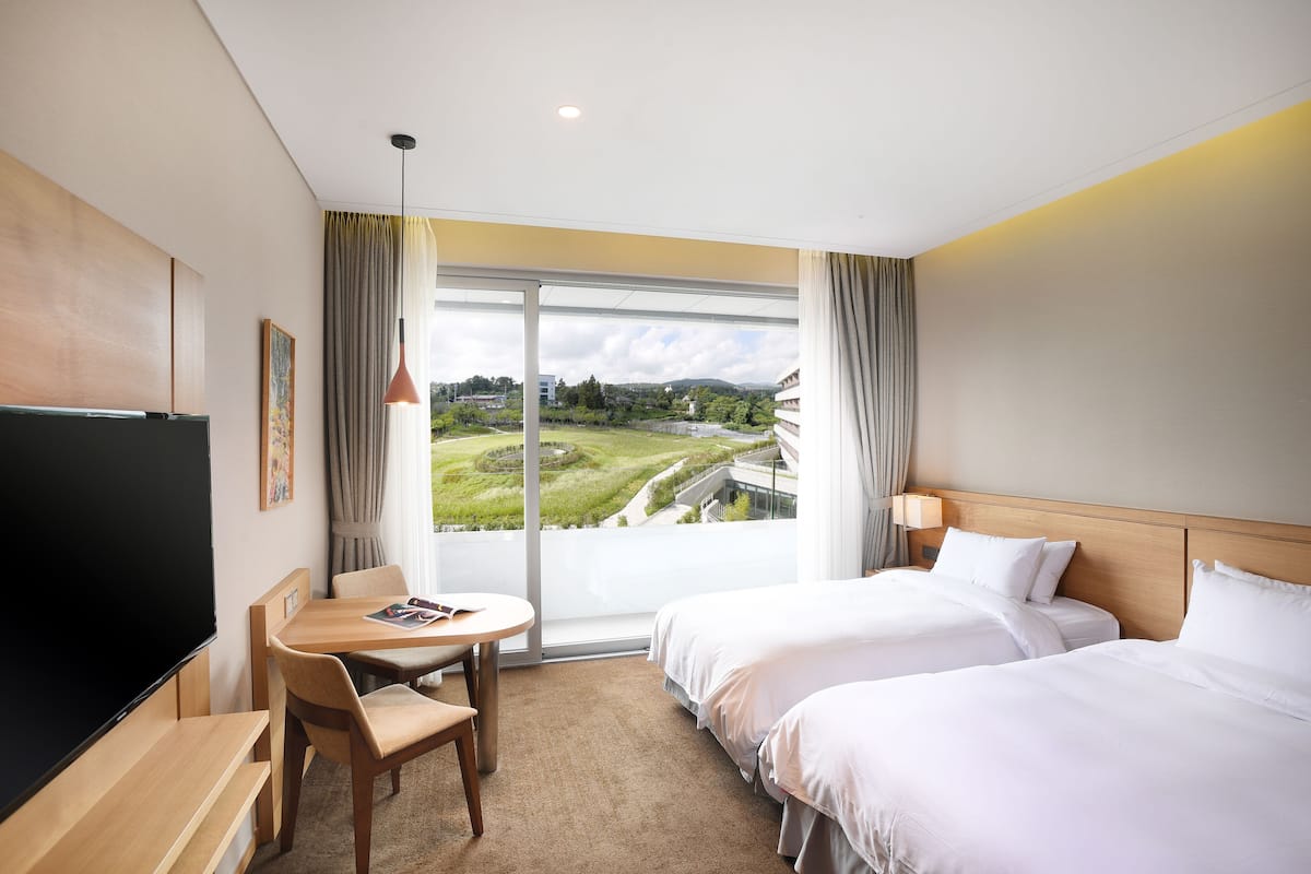 A hotel room with a large TV, a dining table, two beds, and a view of a green landscape through the window.