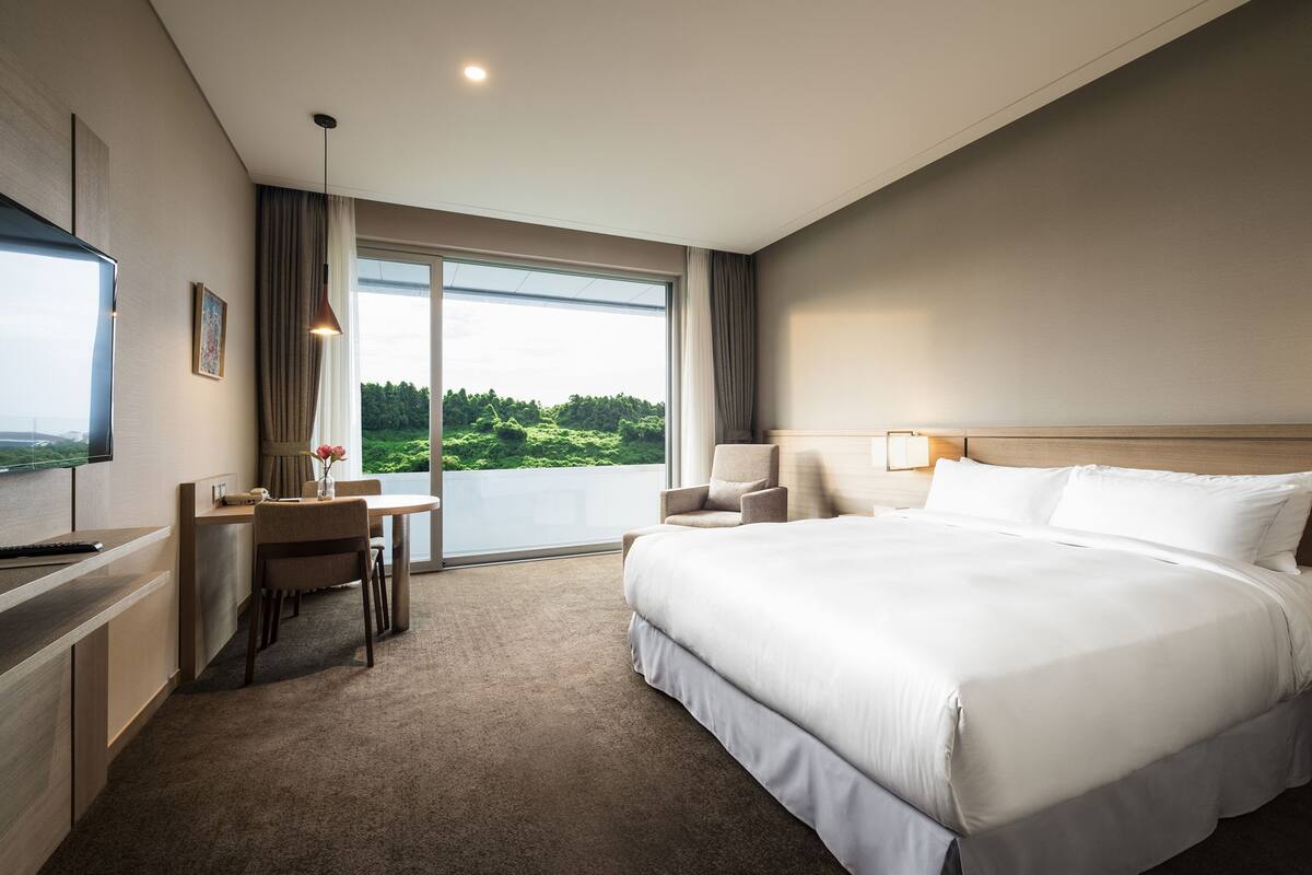 A hotel room with a large bed, a desk with a chair, a television, and a view of greenery through the window.