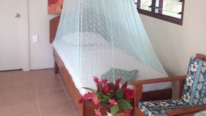 A room with a bed under a mosquito net, a wooden chair with a patterned cushion, a table with a vase of flowers, and a painting on the wall. - Tiavi Mountain Cabins (Siumu)