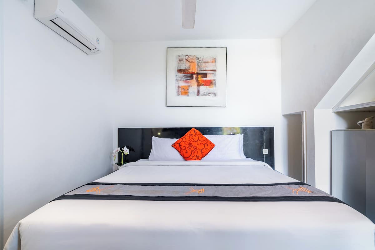A hotel room with a bed, white walls, an air conditioning unit, a framed abstract painting, and a nightstand with a lamp.
