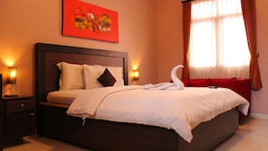 A neatly made bed with a swan-shaped towel, a red painting above it, and two bedside lamps. - Puri Ganesh Hotel (Legian)