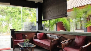 A covered outdoor seating area with wicker furniture and a glass table, overlooking a garden with palm trees. - Puri Ganesh Hotel (Legian)