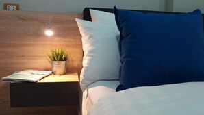 Un lit bien fait, avec des oreillers blancs et bleus, une petite table de chevet ornée d’une plante et d’un livre, et une prise électrique au-dessus de la table.