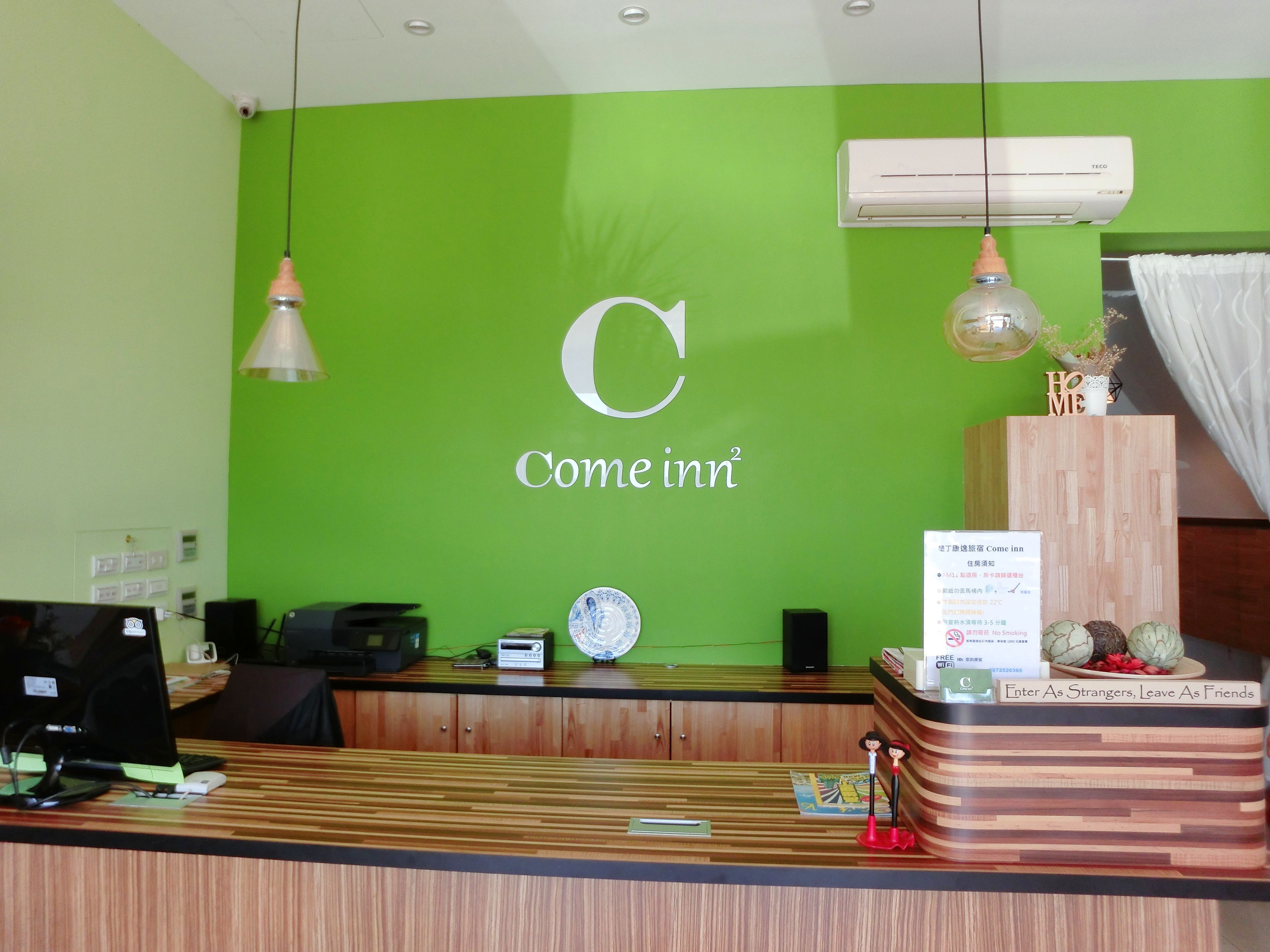 A hotel reception desk with a green wall, a large 'C' logo, and a sign that reads 'Come inn'. There is a wooden counter, a computer, and a small decorative figure.