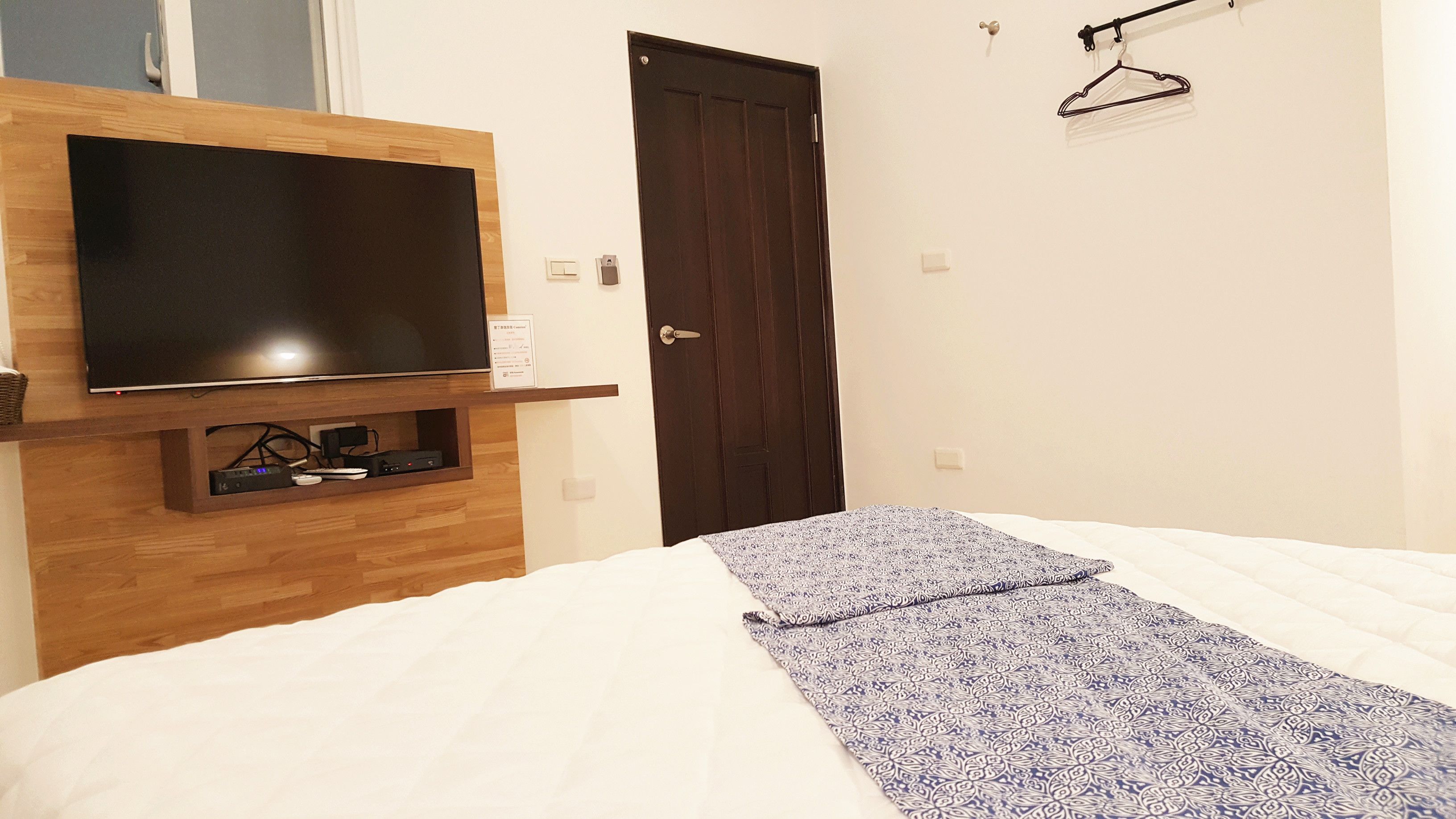 Un dormitorio con cama, televisión montada en un estante de madera, una puerta y un perchero en la pared.
