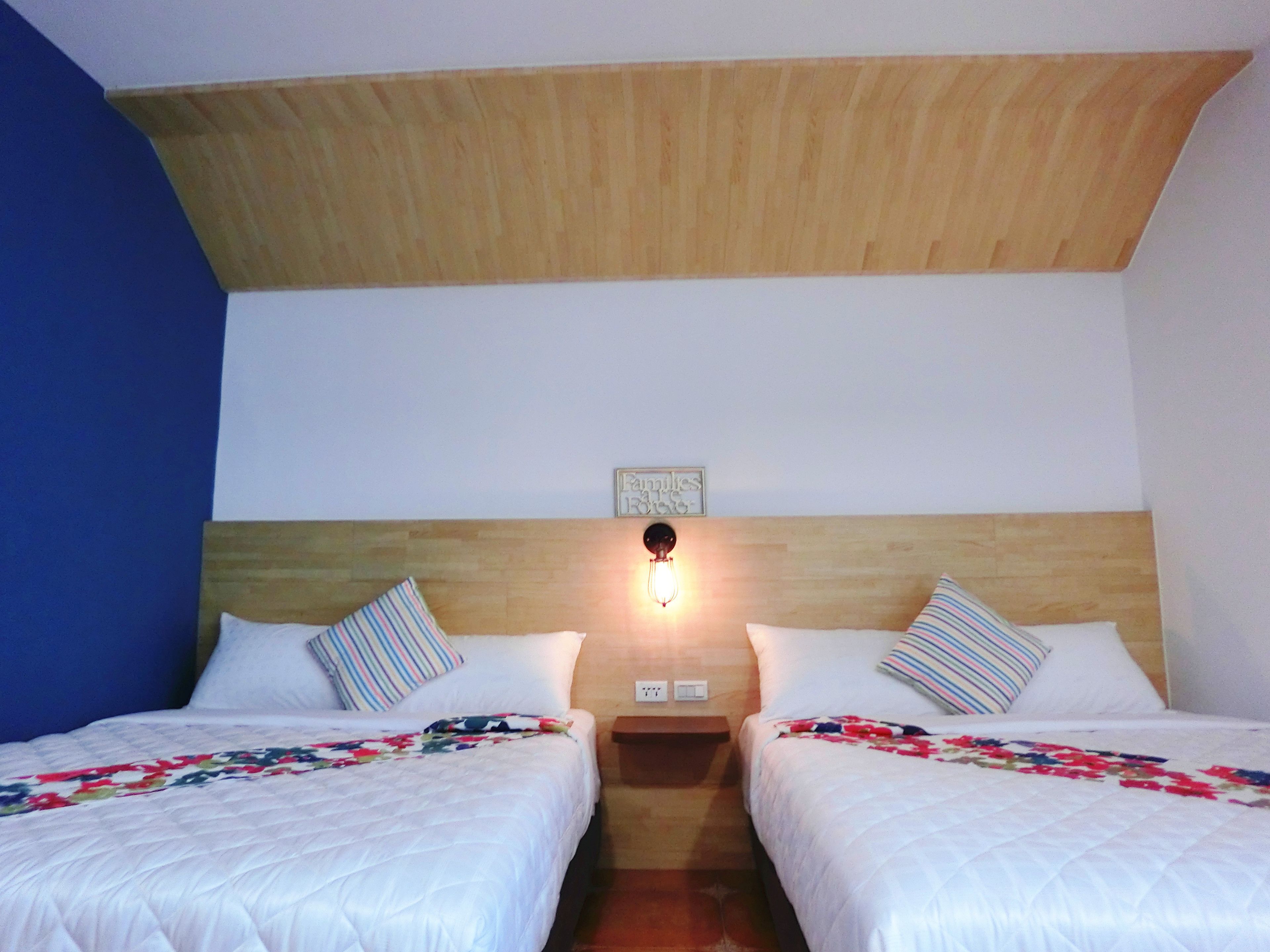 A room with two beds, each with a floral-patterned bedspread, a wooden headboard, and a wall-mounted light fixture.