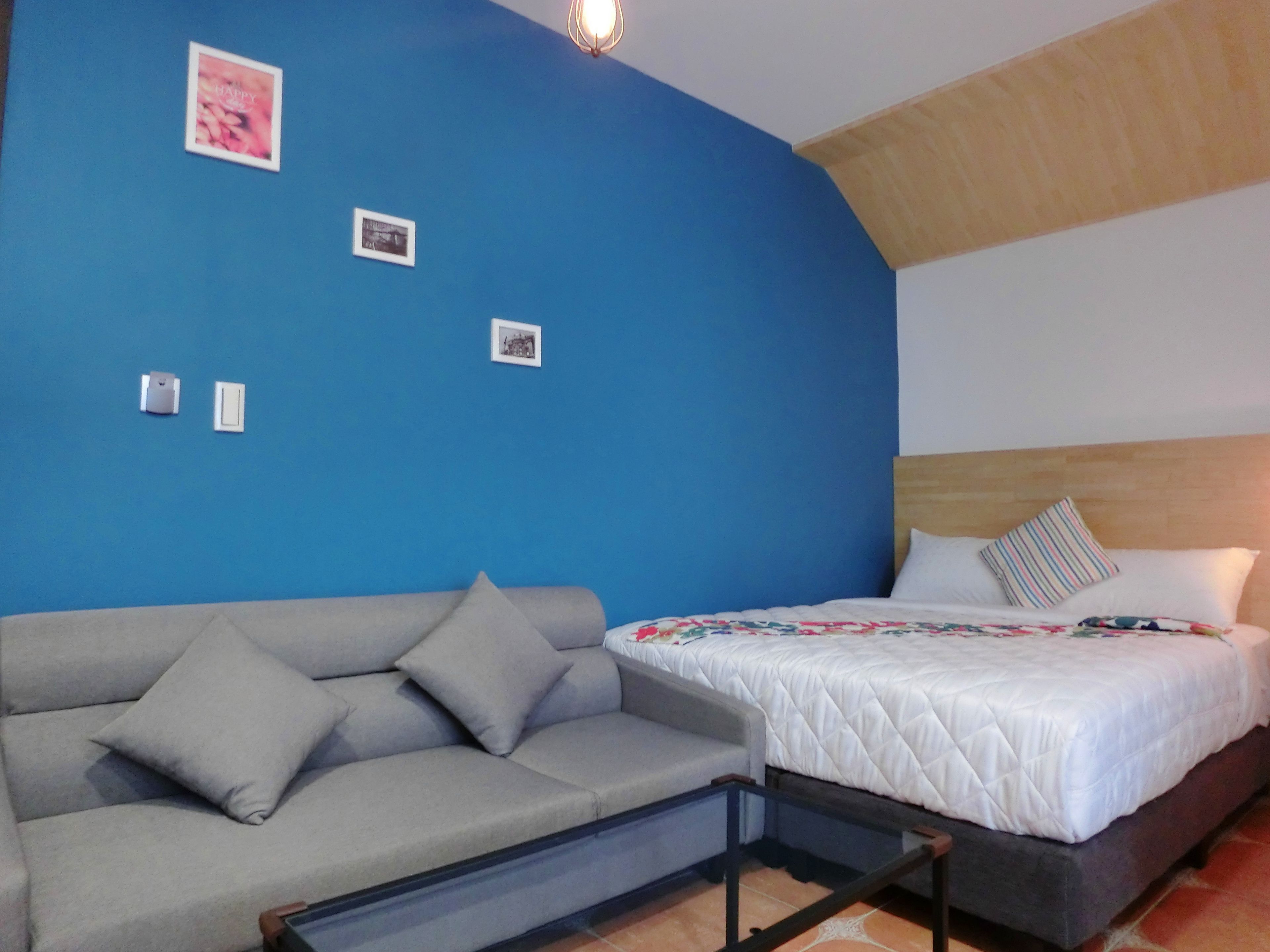 Une pièce avec un mur d’accent bleu, un canapé gris, un lit avec une literie blanche et une tête de lit en bois.