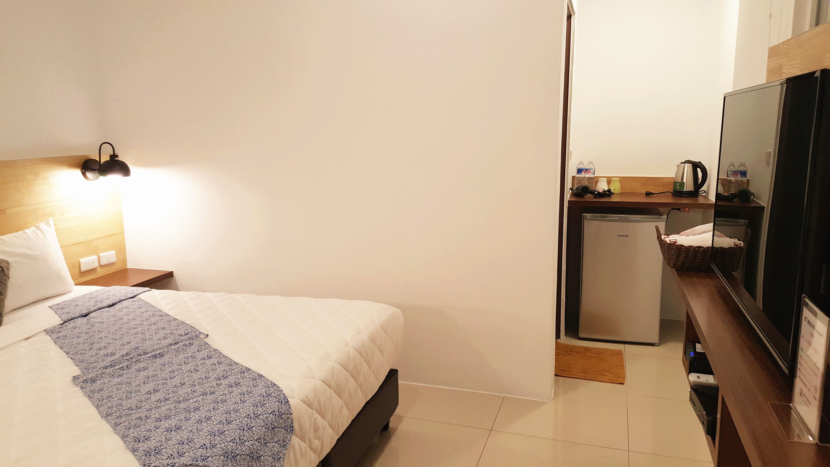A hotel room with a bed, a nightstand, a wall-mounted lamp, a small kitchenette with a refrigerator, and a mirror.