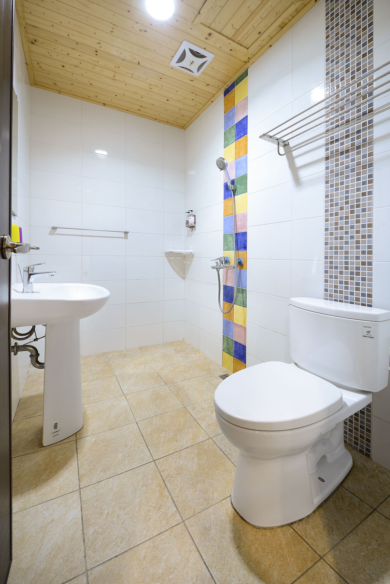 A bathroom with a wooden ceiling, white tiles, a colorful vertical tile accent, a white sink, a toilet, and a shower head.