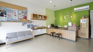Zona de recepción de hotel con una pared verde, mostrador de madera, un sofá gris y un tablón de anuncios con varios avisos.