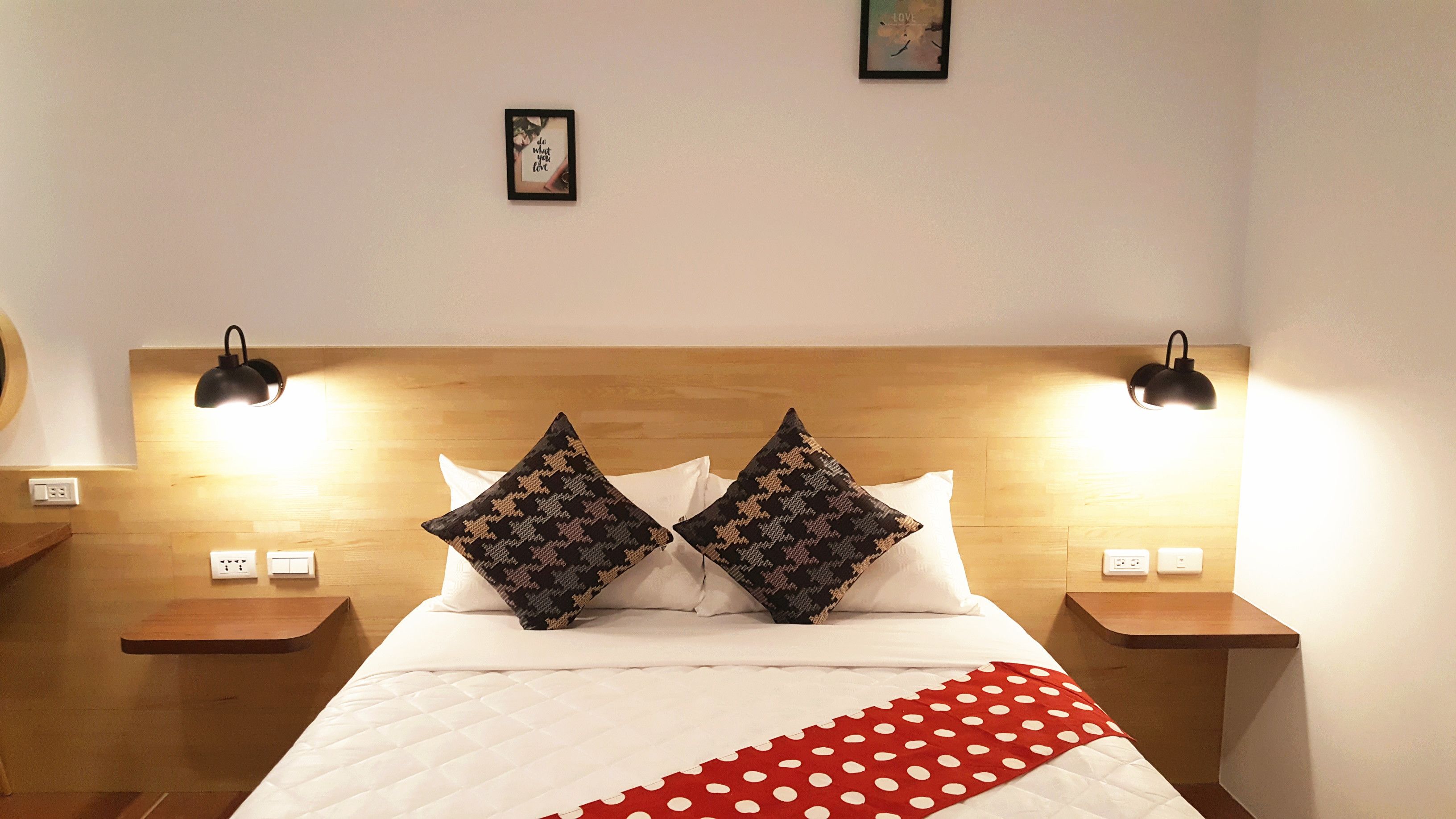 A neatly made bed with white bedding and two patterned pillows, flanked by two bedside tables with lamps, in a room with a wooden headboard and a small framed picture on the wall.