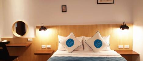 Habitación de hotel con cabecera de madera, dos lámparas de noche, un espejo redondo y dos almohadas con frases motivacionales.