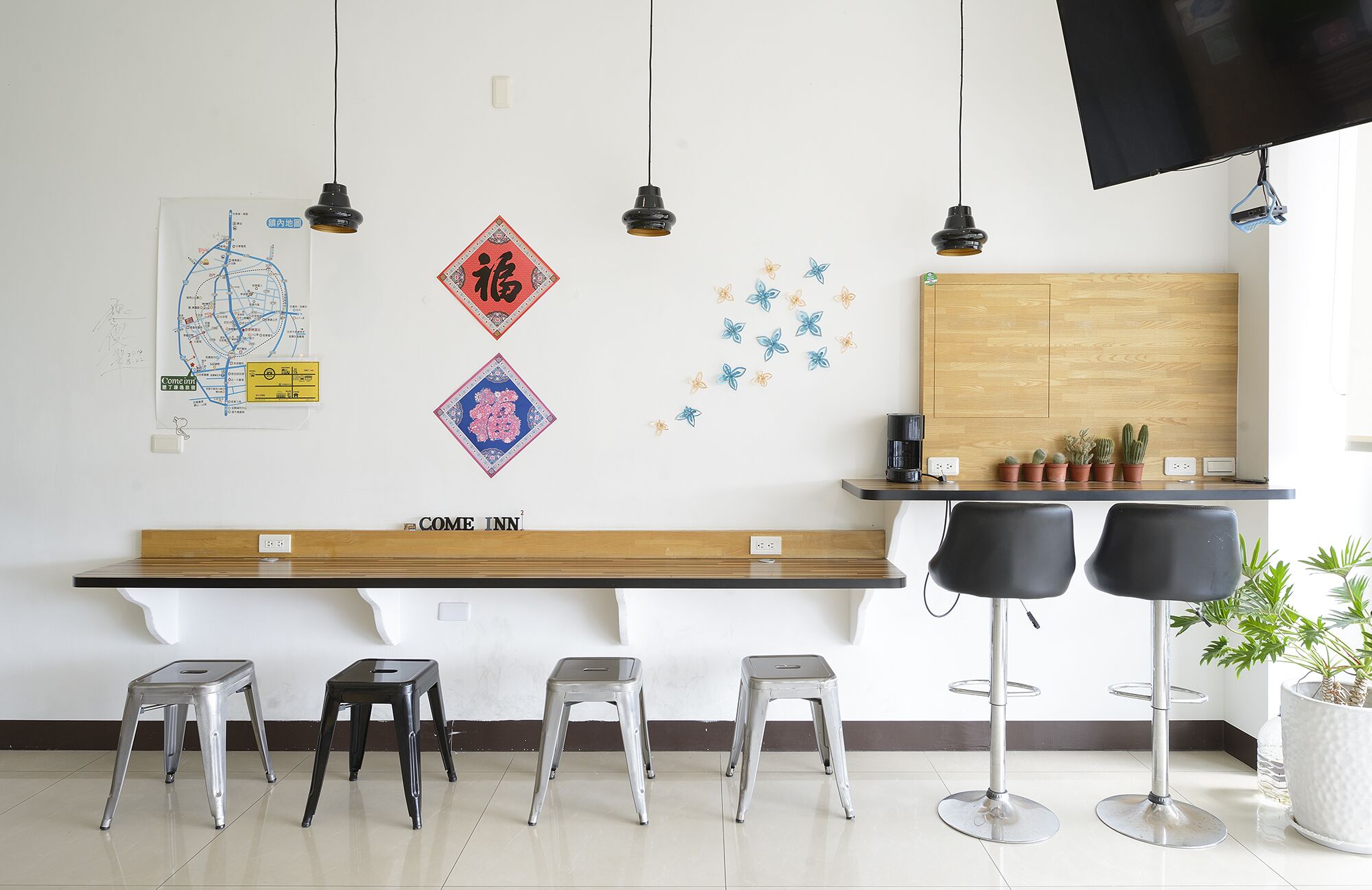 Un espace d’accueil d’hôtel avec un comptoir en bois, deux tabourets de bar noirs, une carte, des éléments décoratifs suspendus au mur et un téléviseur fixé au mur.