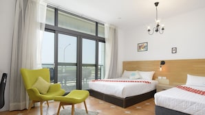 Una habitación de hotel con un ventanal grande, una cama con ropa blanca, un sillón verde y un candelabro.
