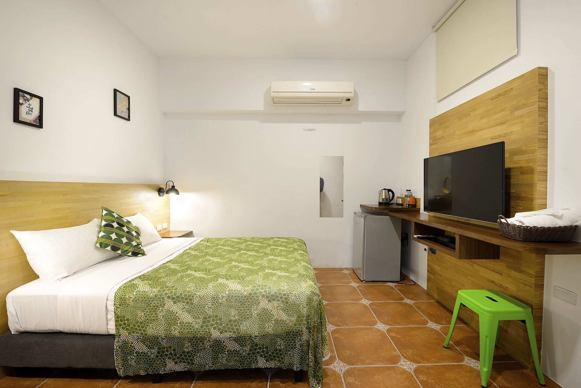Una camera d'albergo con un letto, una scrivania con televisore, un angolo cottura e uno sgabello verde.