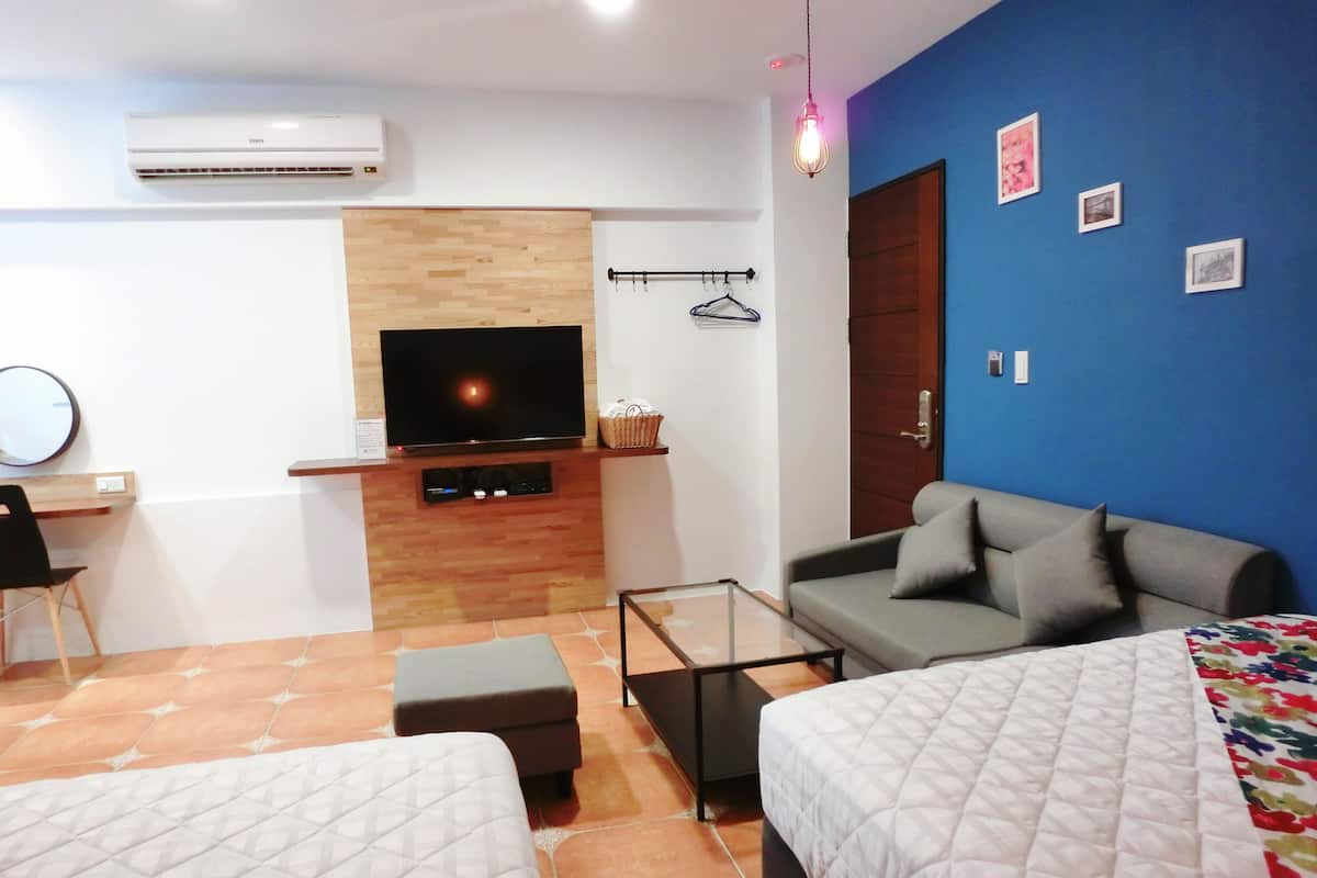 A modern hotel room with a flat-screen TV, a sofa, two beds, a small table, and a hanging light fixture.