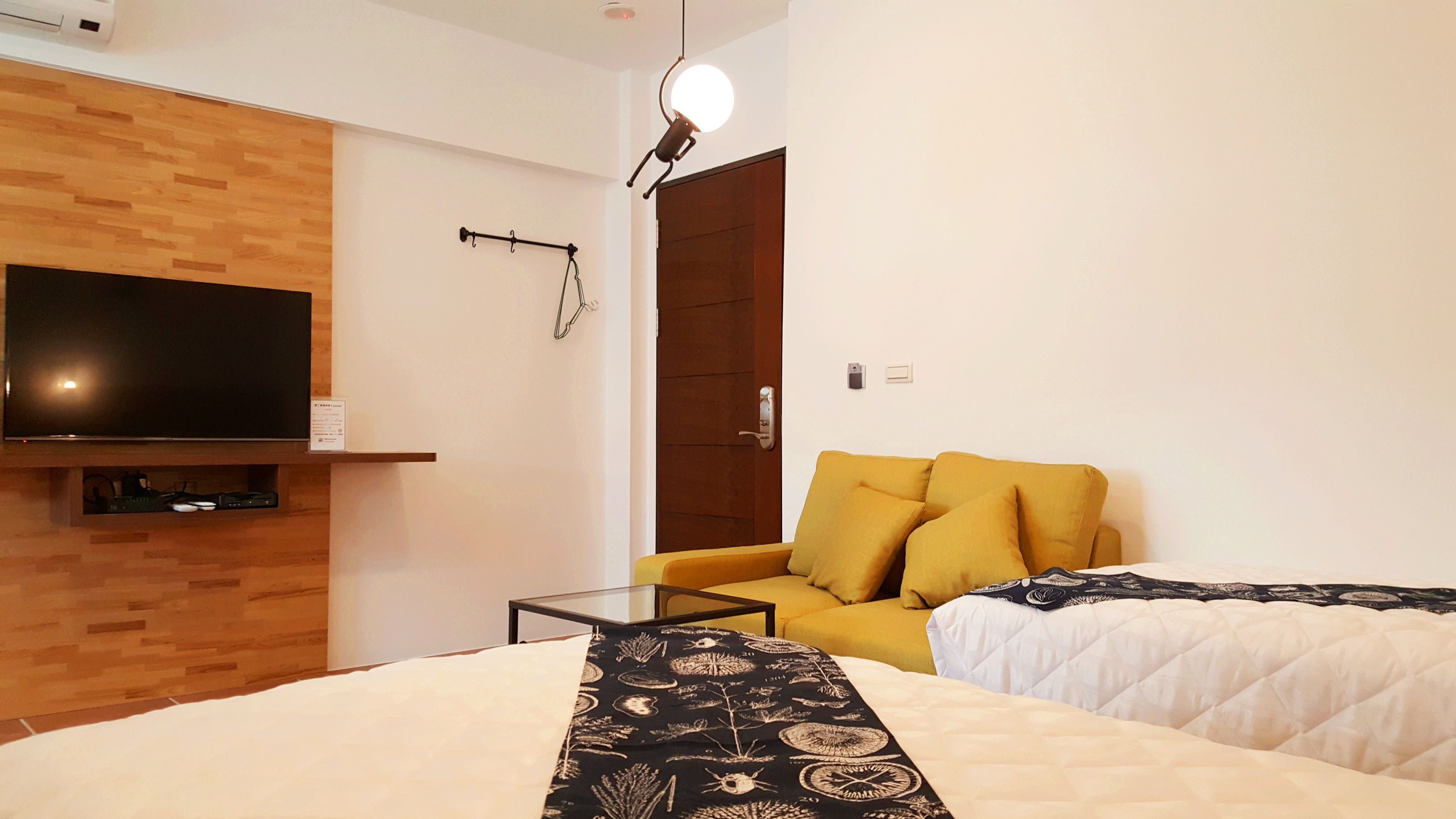 Quarto de hotel com parede de madeira, TV de tela plana, sofá amarelo, duas camas com colchas estampadas e uma pequena mesa.