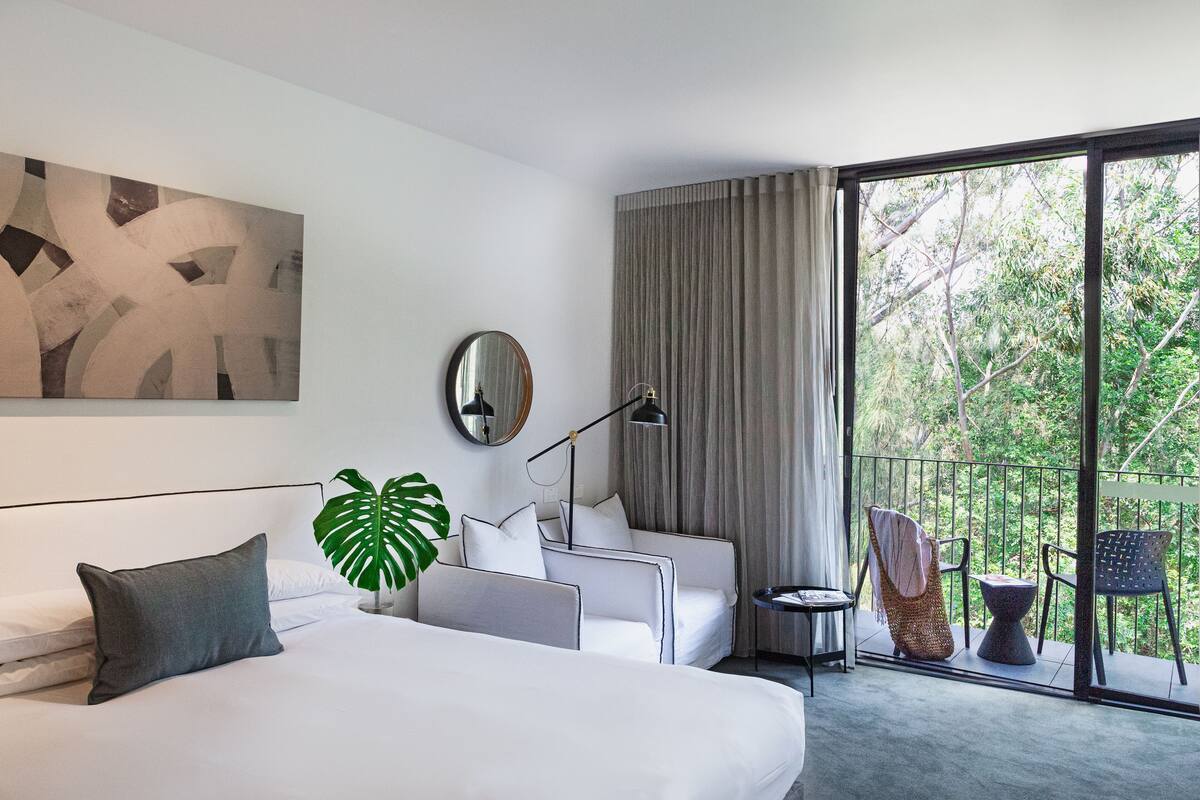 Una habitación de hotel moderna con una cama grande, un sofá, una mesita y un balcón con vistas a árboles.