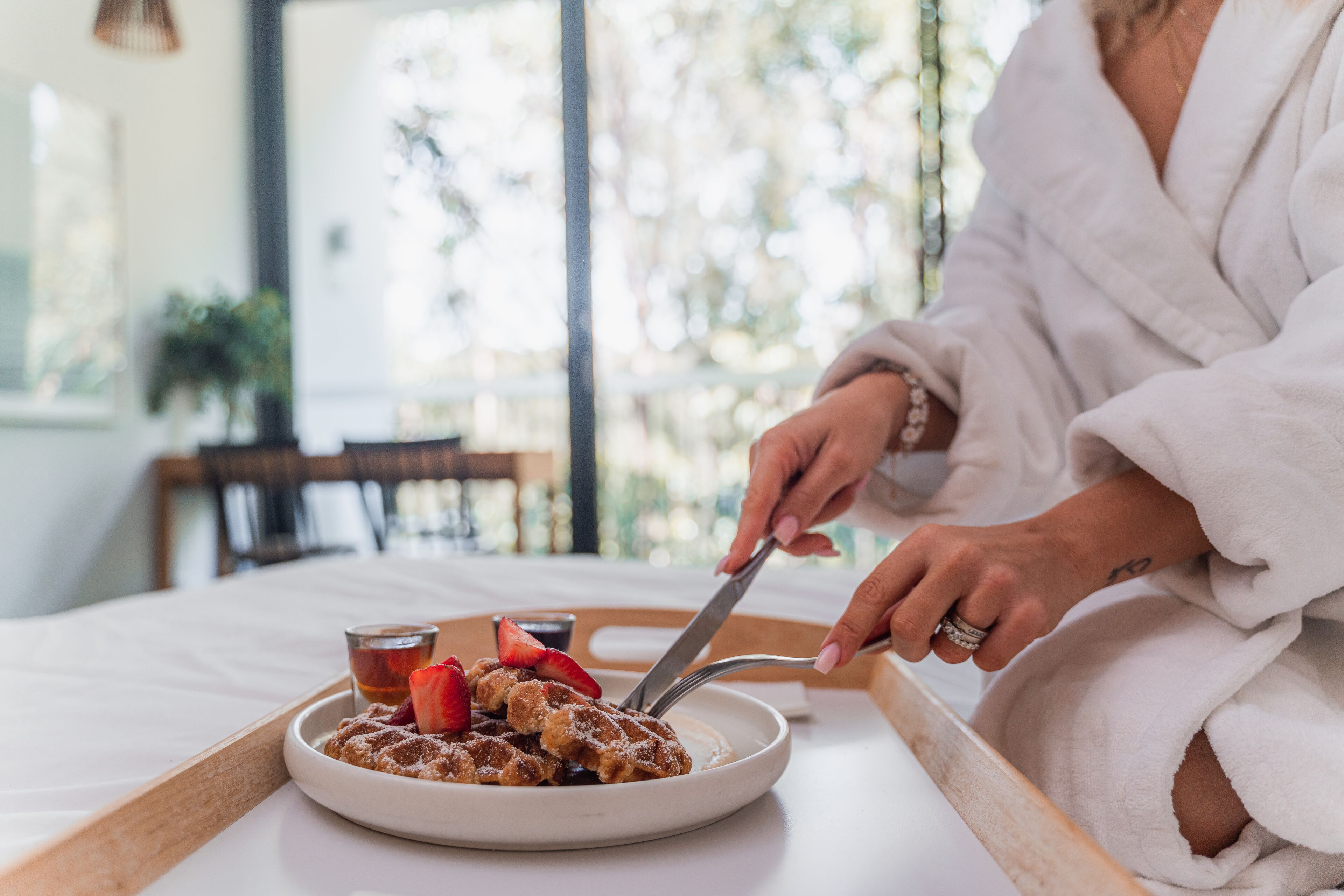 A person in a white robe eating waffles with strawberries and syrup on a tray.
