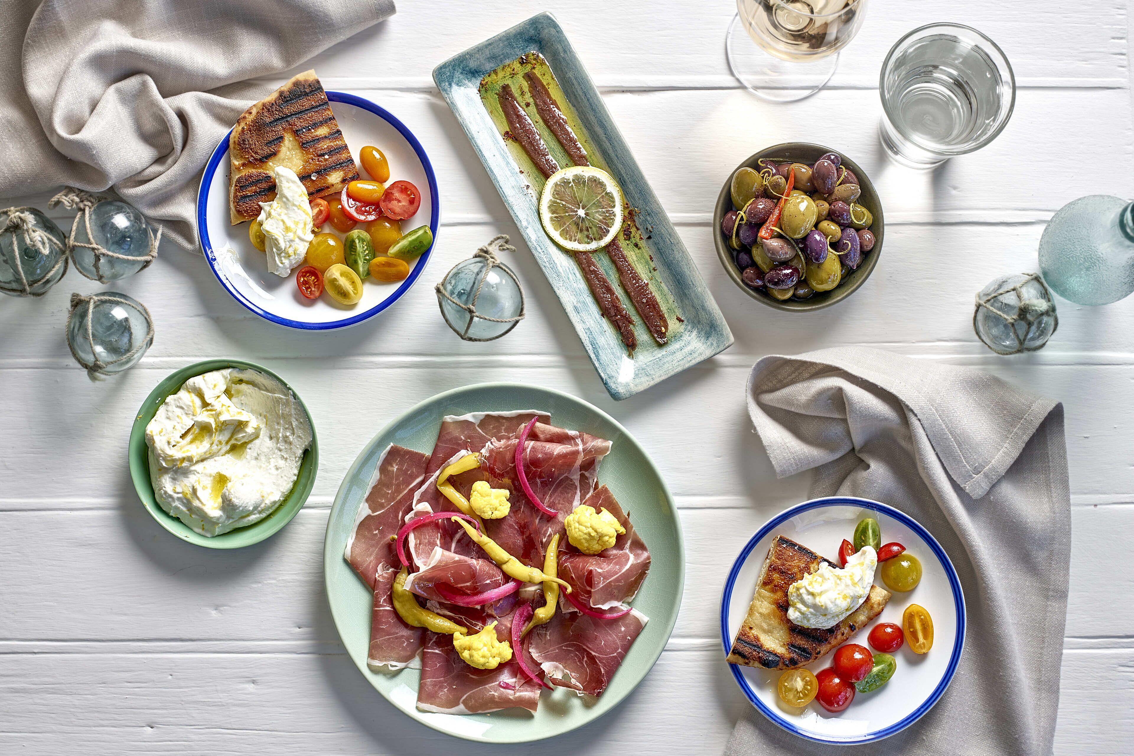 Una mesa con diversos platos que incluyen carne a la parrilla, queso y verduras, acompañada de bebidas y cristalería decorativa.