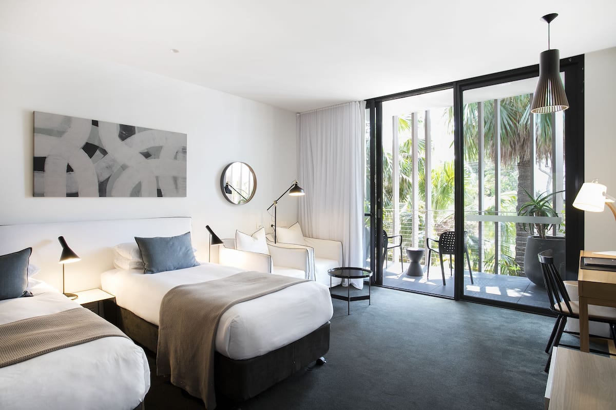 Una habitación de hotel moderna con dos camas, un escritorio, una silla, una mesita, una lámpara, un espejo y un amplio ventanal con vistas a zonas verdes.
