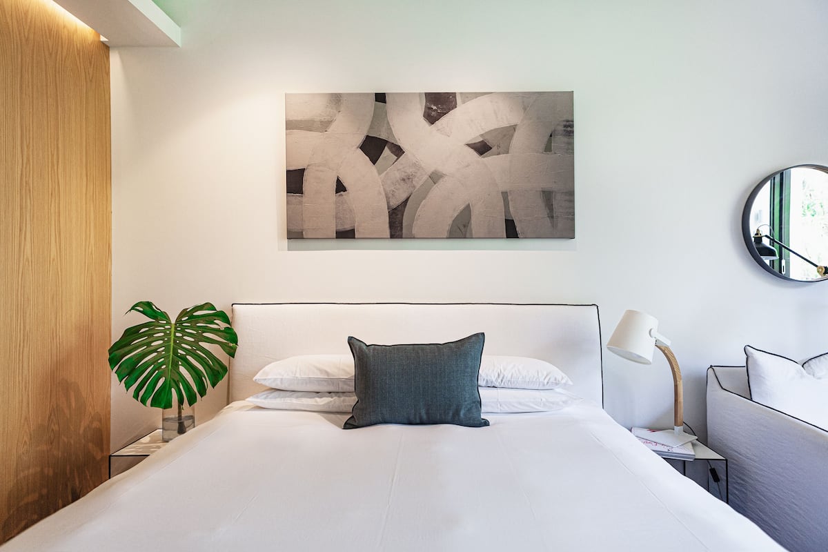 Una habitación de hotel moderna con una cama grande, una mesita de noche con lámpara, una planta y un cuadro abstracto colgado en la pared.