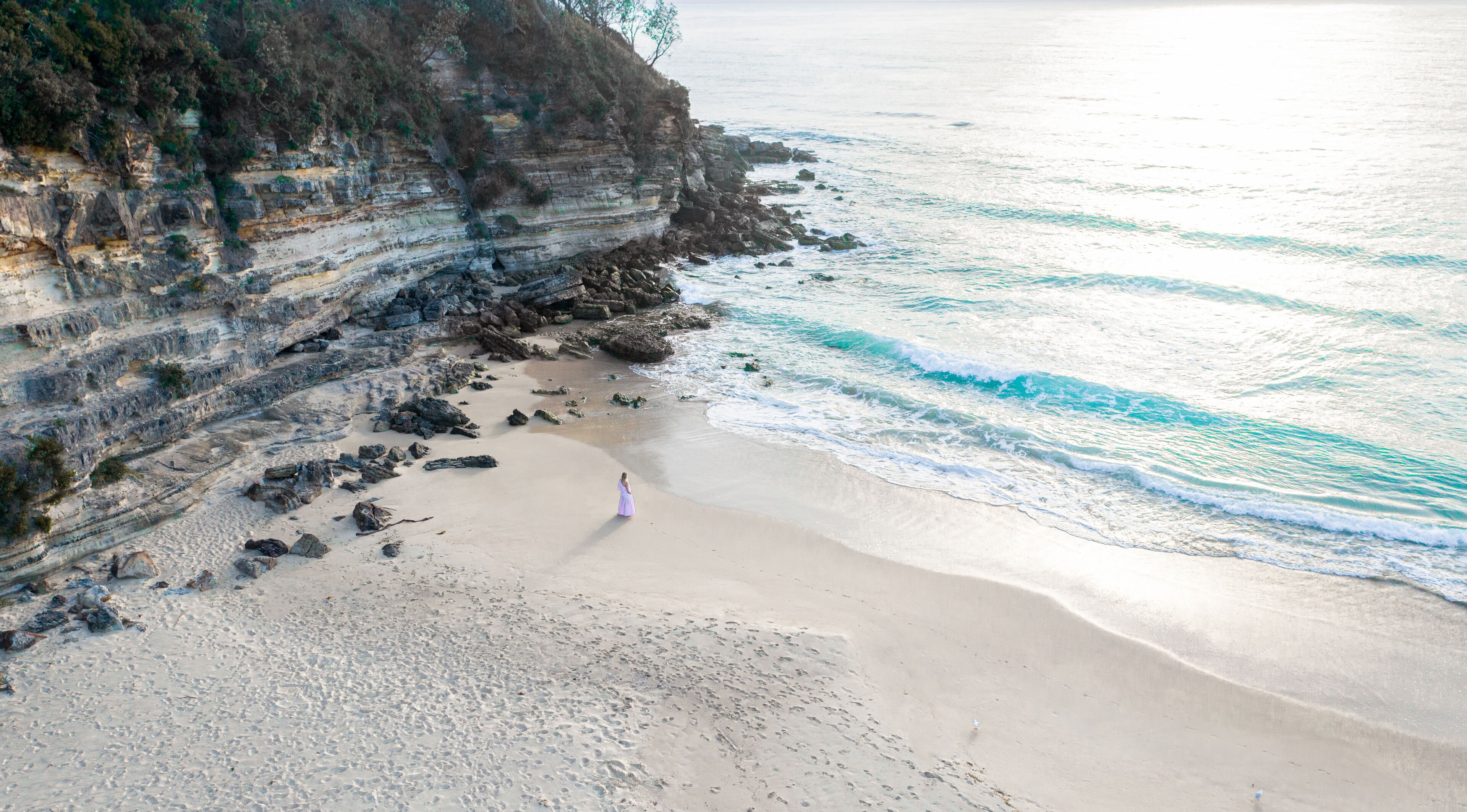 Une plage avec une falaise rocheuse et une personne vêtue d’une robe violette qui marche sur le sable.