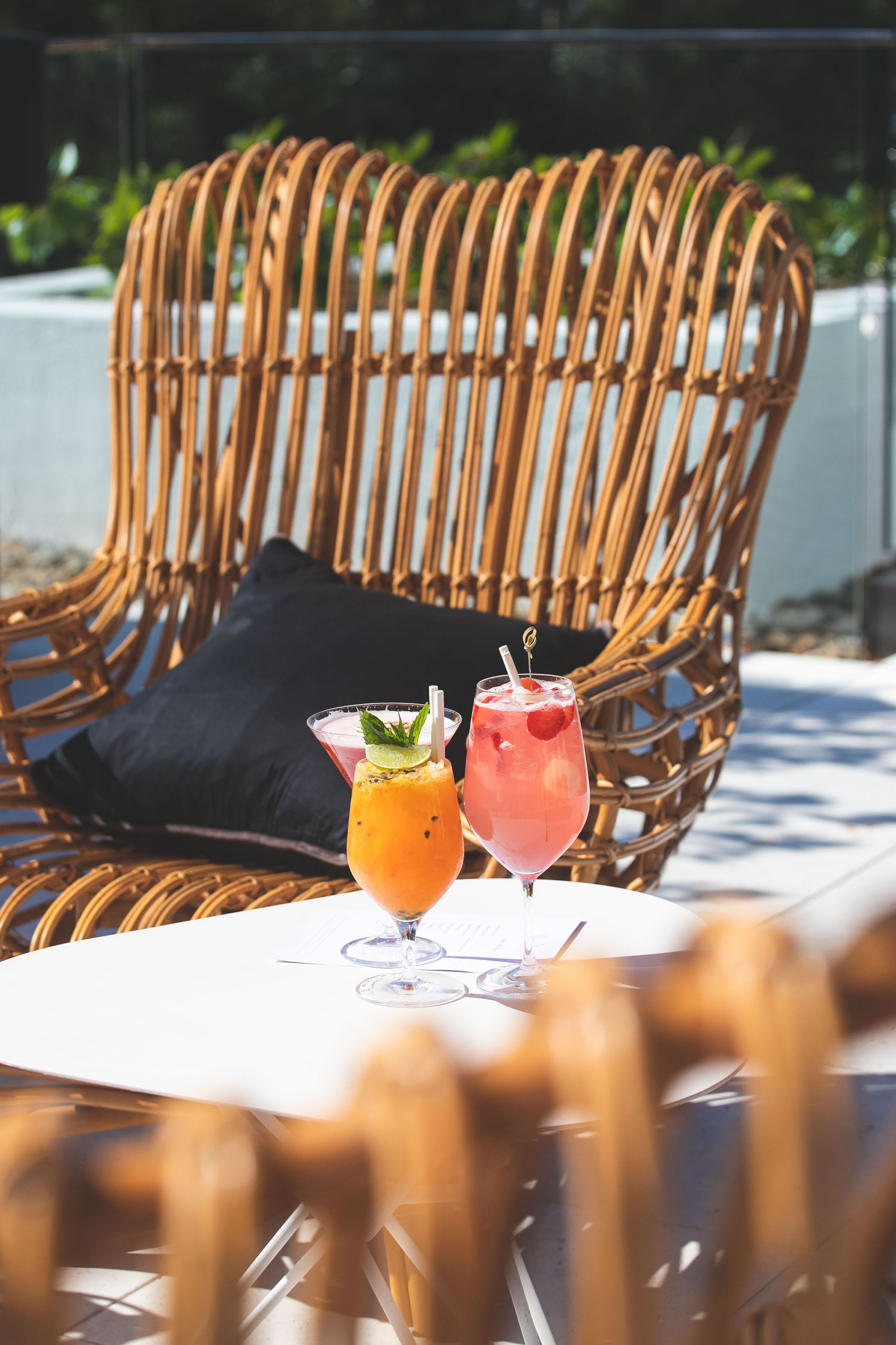 A wicker chair with a black cushion, two drinks on a table, and a clear view of the outdoors.
