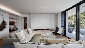 Una sala de estar moderna con un sofá, una mesa de centro y una chimenea. Hay una estantería y un ventanal con vistas al exterior.