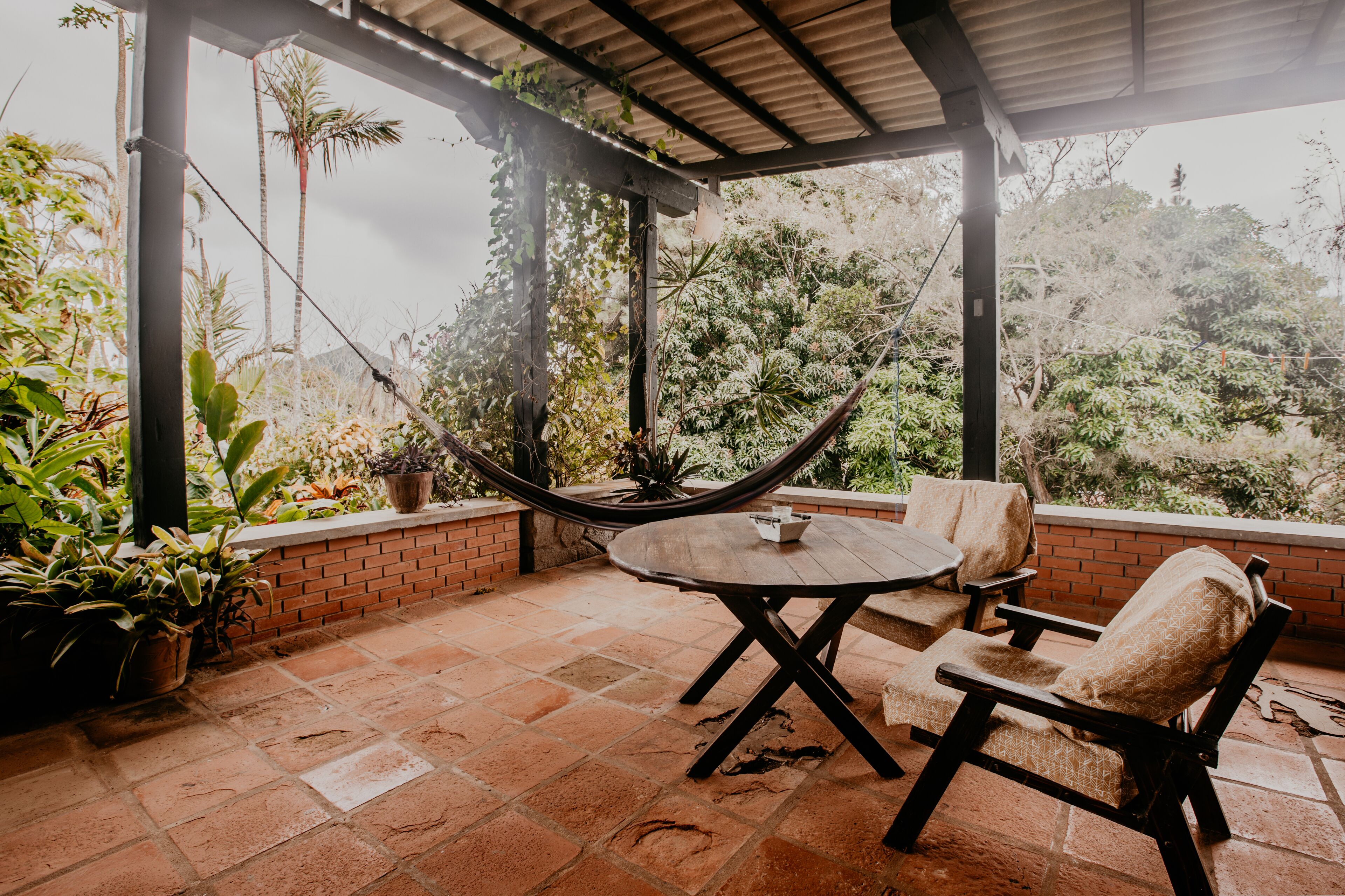 A covered patio with a round table and two cushioned chairs, surrounded by potted plants and a hammock.