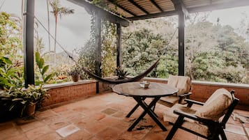 A covered patio with a round table and two cushioned chairs, surrounded by potted plants and a hammock.