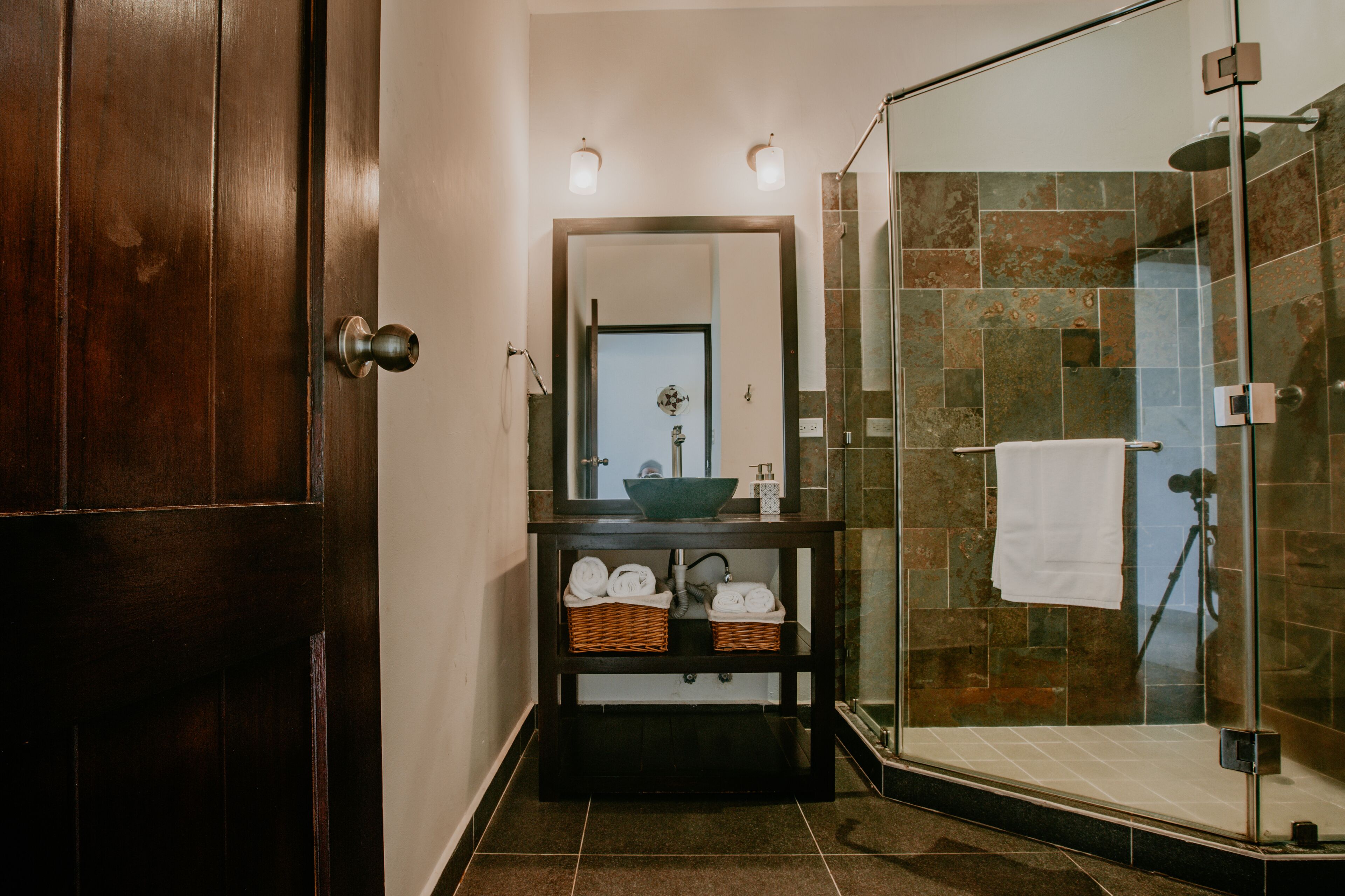 A bathroom with a wooden door, a mirror above a sink with baskets, and a glass shower enclosure.