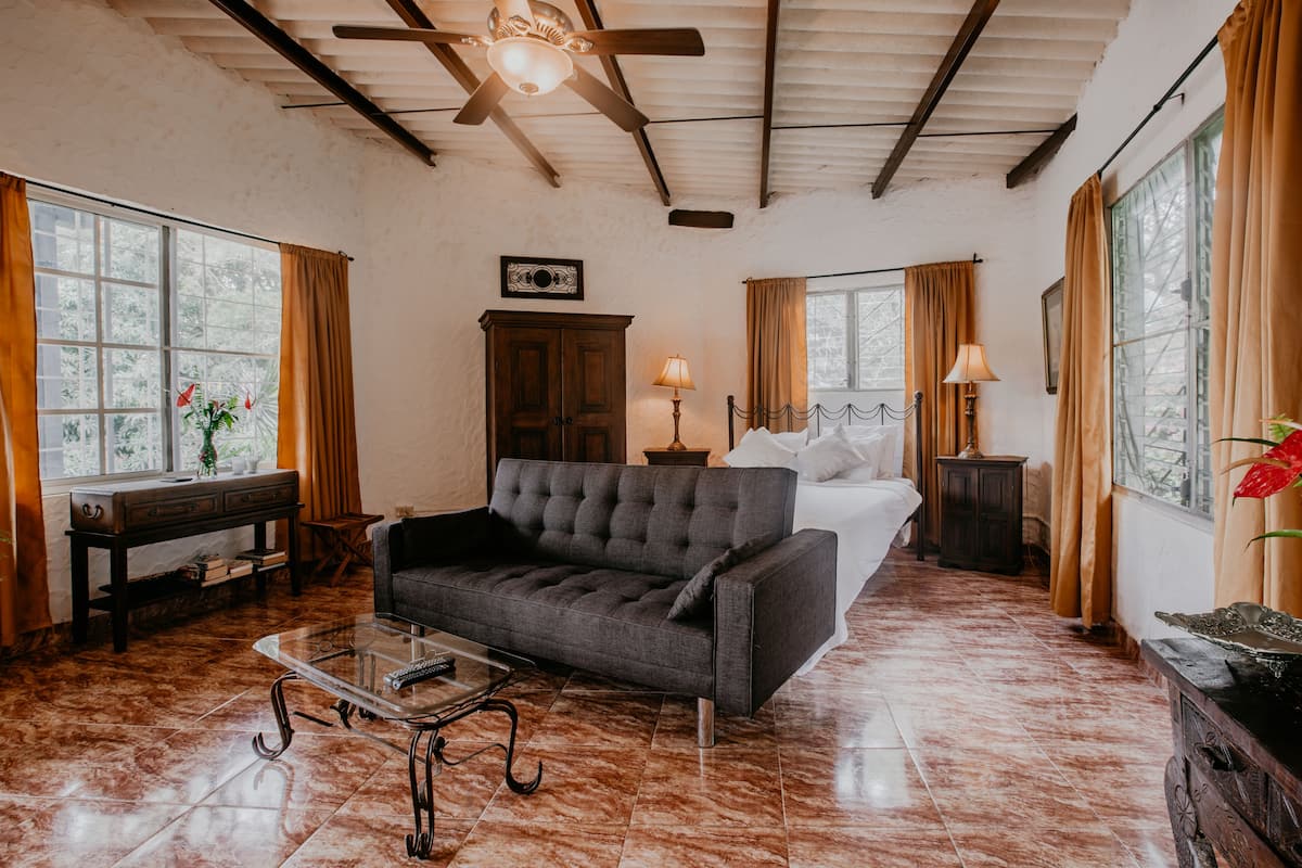 A spacious room with a large bed, a sofa, a small table, and a ceiling fan.