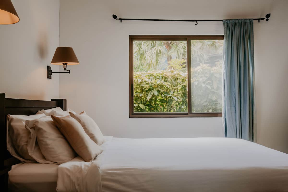 A bedroom with a bed, pillows, a nightstand, a wall-mounted lamp, a window with curtains, and a view of greenery outside.