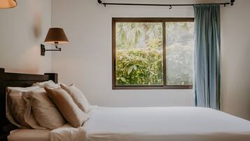 Un dormitorio con cama, almohadas, una mesita de noche, una lámpara de pared, una ventana con cortinas y vistas a zonas verdes.
