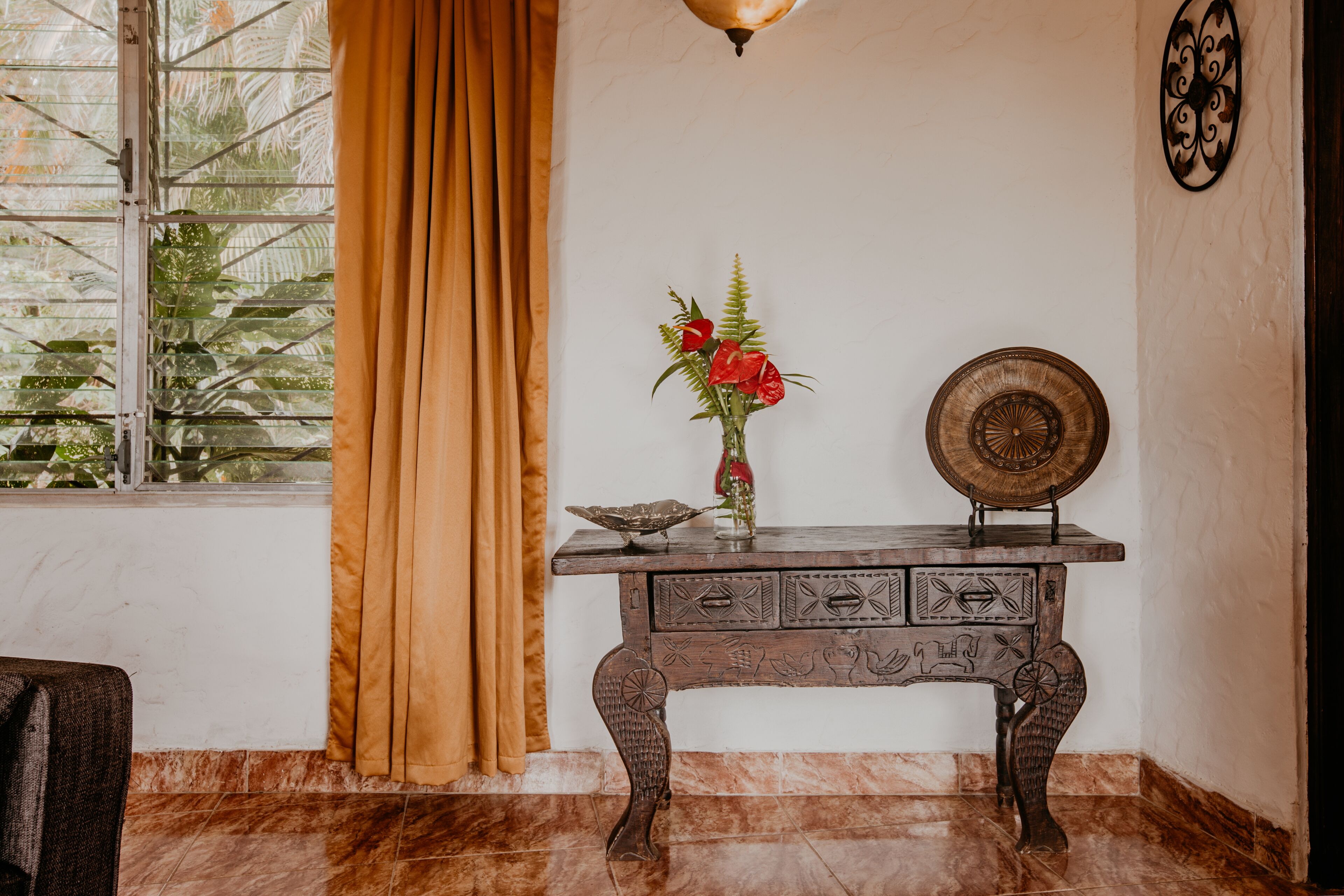 Um quarto com uma mesa consola de madeira, um vaso com flores vermelhas, um prato decorativo e uma janela com cortinas.