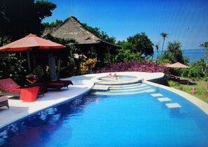 A swimming pool with lounge chairs and umbrellas, surrounded by tropical plants and a view of the sea. - TOYABALI - Resort (Tulamben)