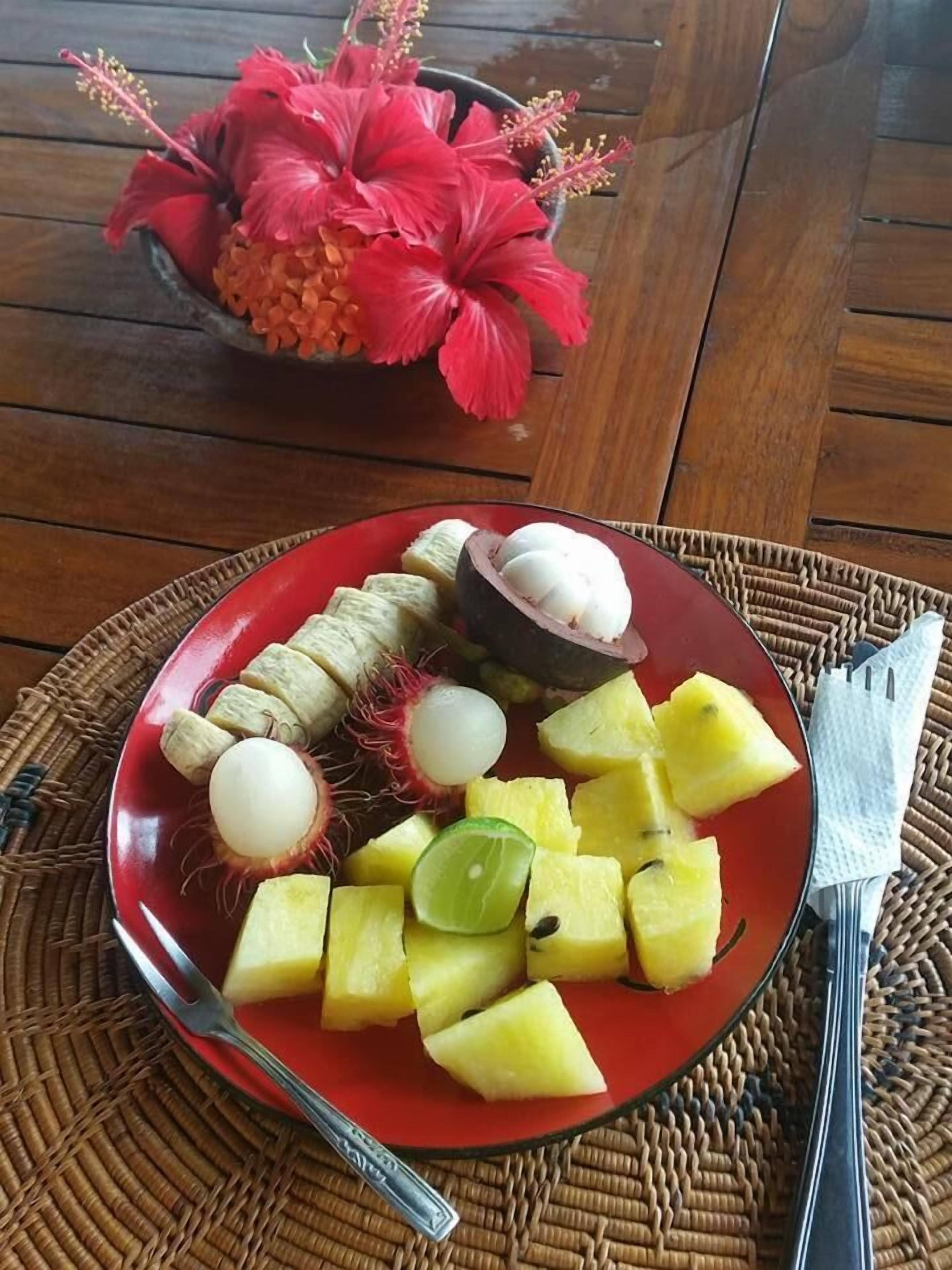 A plate of fruit including pineapple, rambutan, and lychee, with a red bowl of flowers in the background.