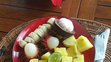 A plate of fruit including pineapple, rambutan, and lychee, with a red bowl of flowers in the background.
