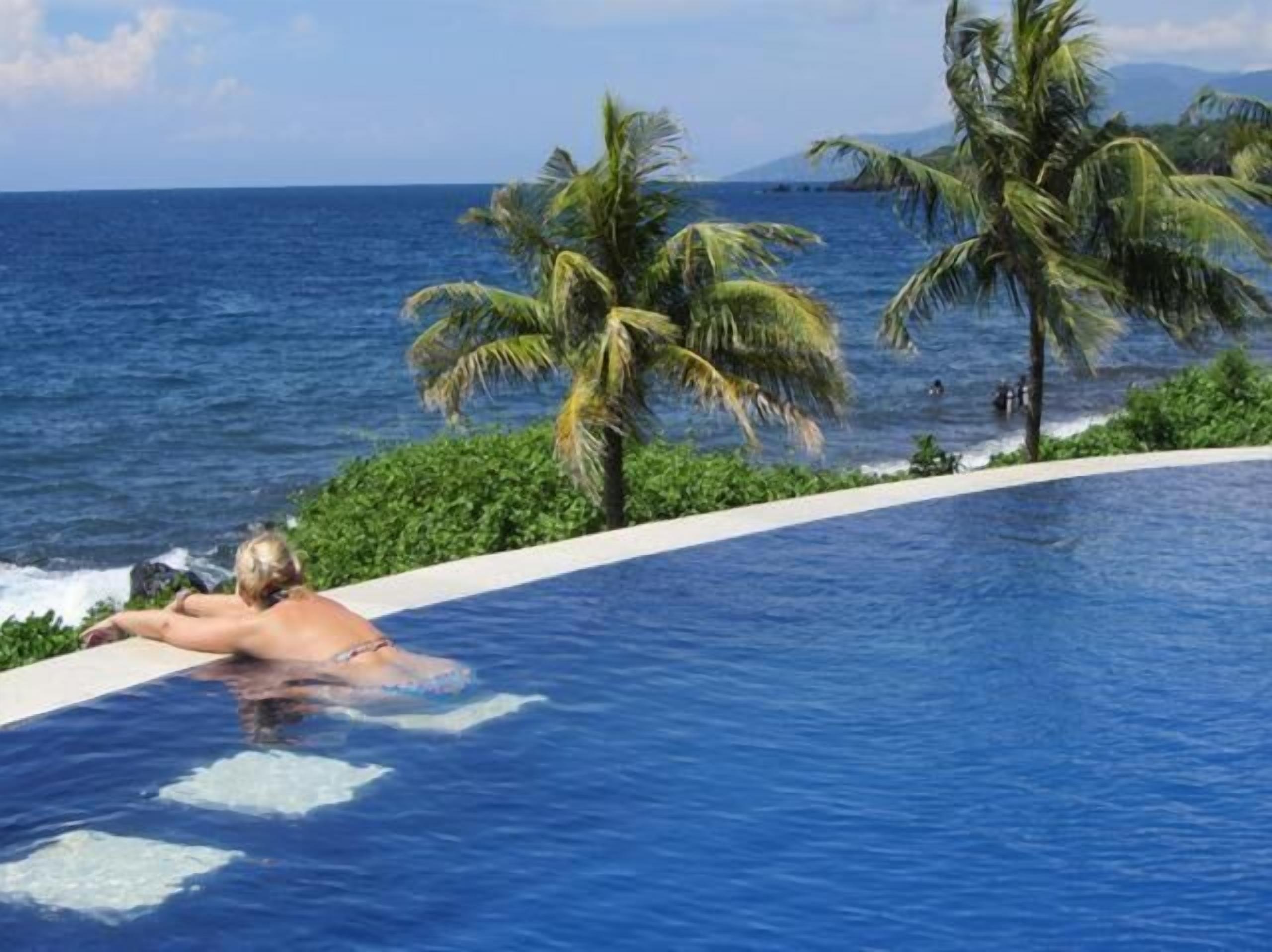 Una persona relajándose en una piscina con vista al océano y palmeras.