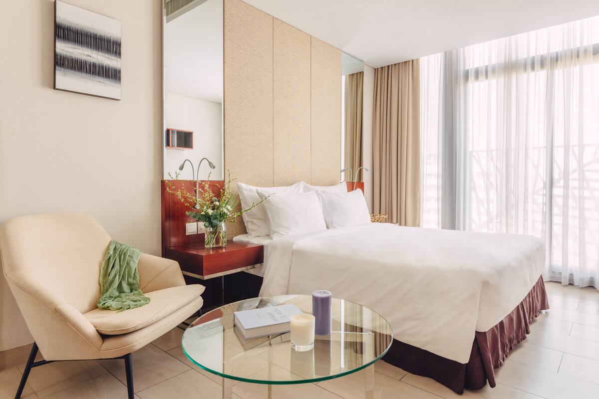 Een hotelkamer met een bed, een stoel, een glazen bijzettafeltje, een nachtkastje met een lamp en een groot raam met gordijnen.