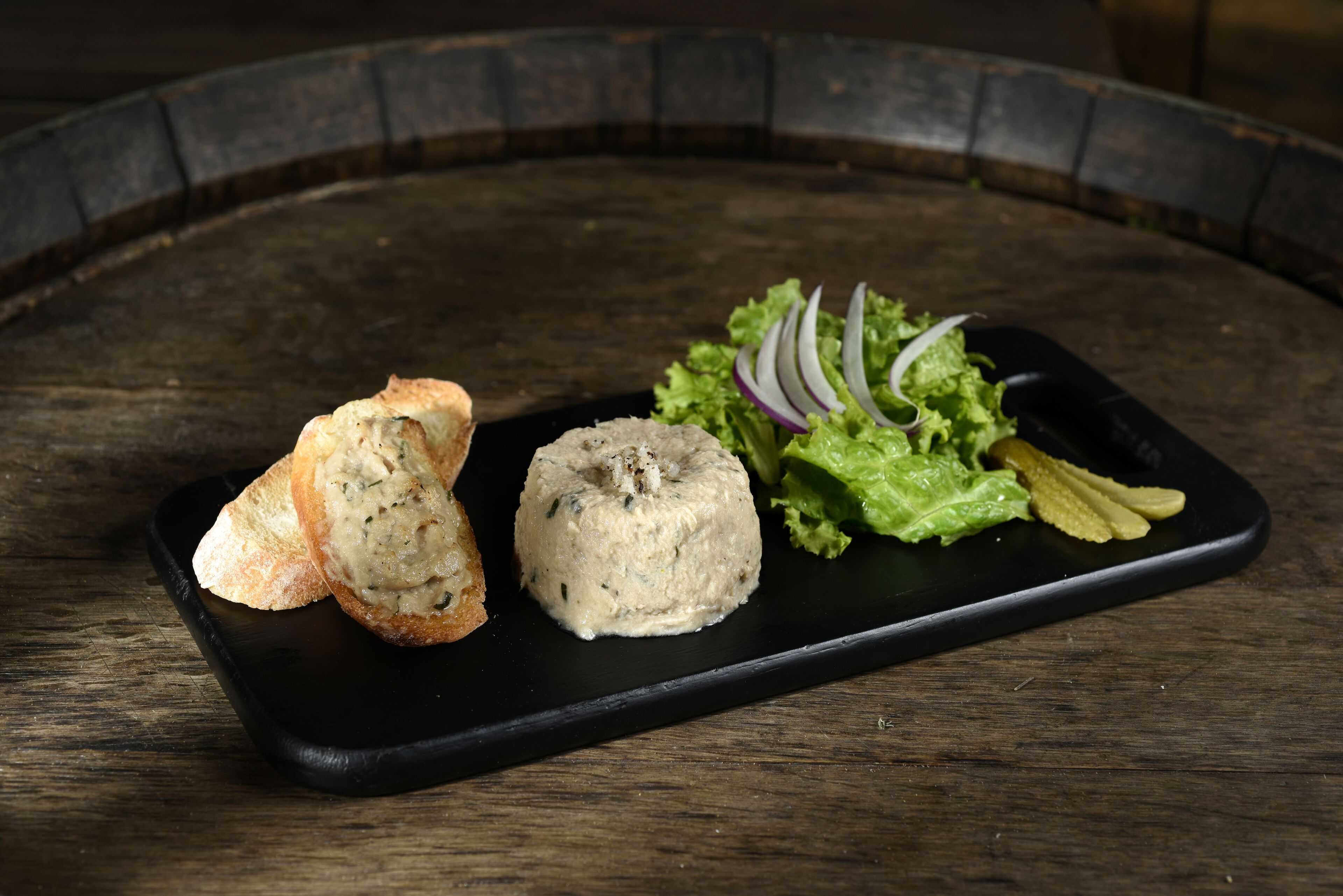 A black rectangular plate with a piece of bread and a round food item, accompanied by a small salad with lettuce and sliced onions on a wooden table.