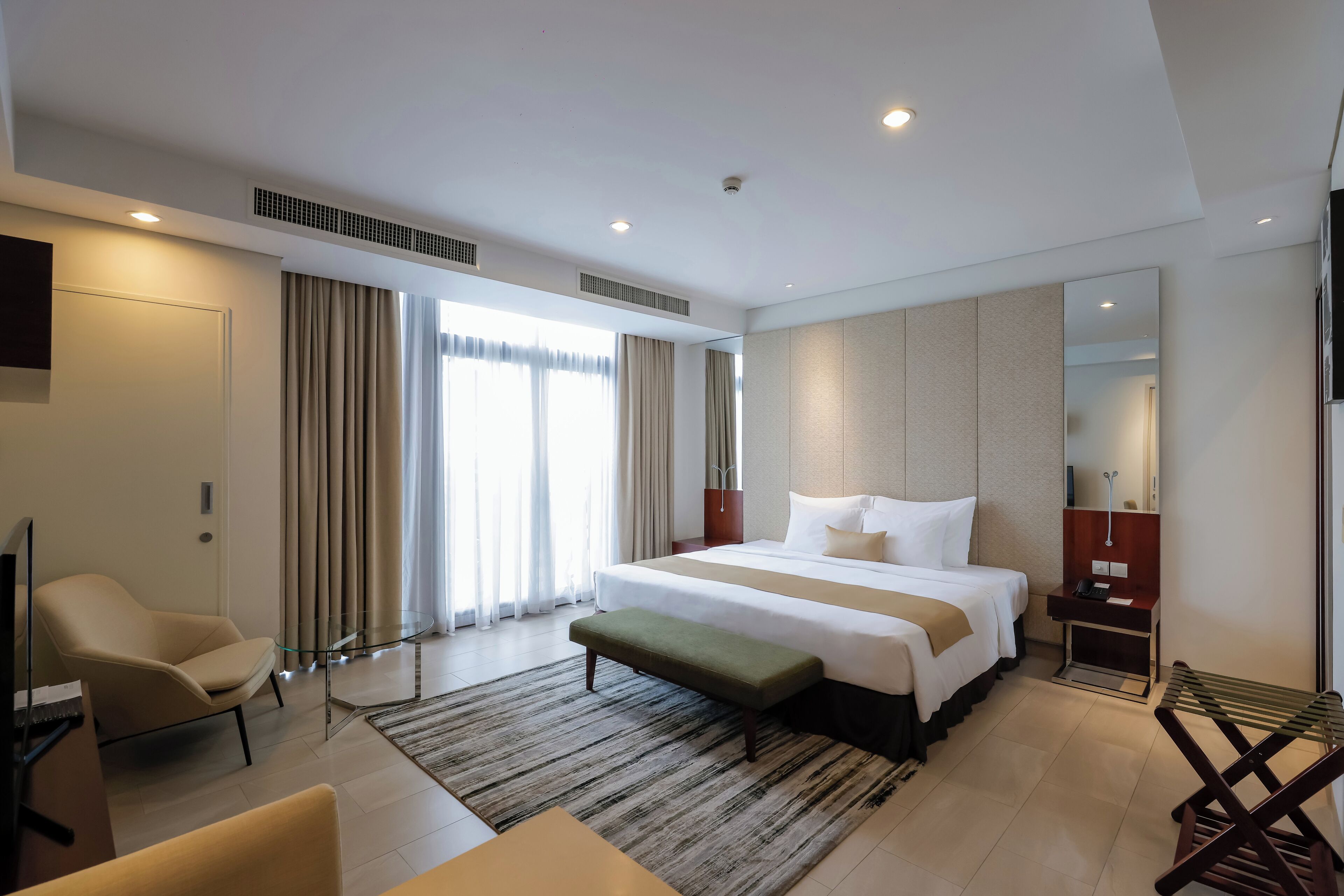 A modern hotel room with a large bed, a sitting area with a chair, a small table, and a desk with a telephone.